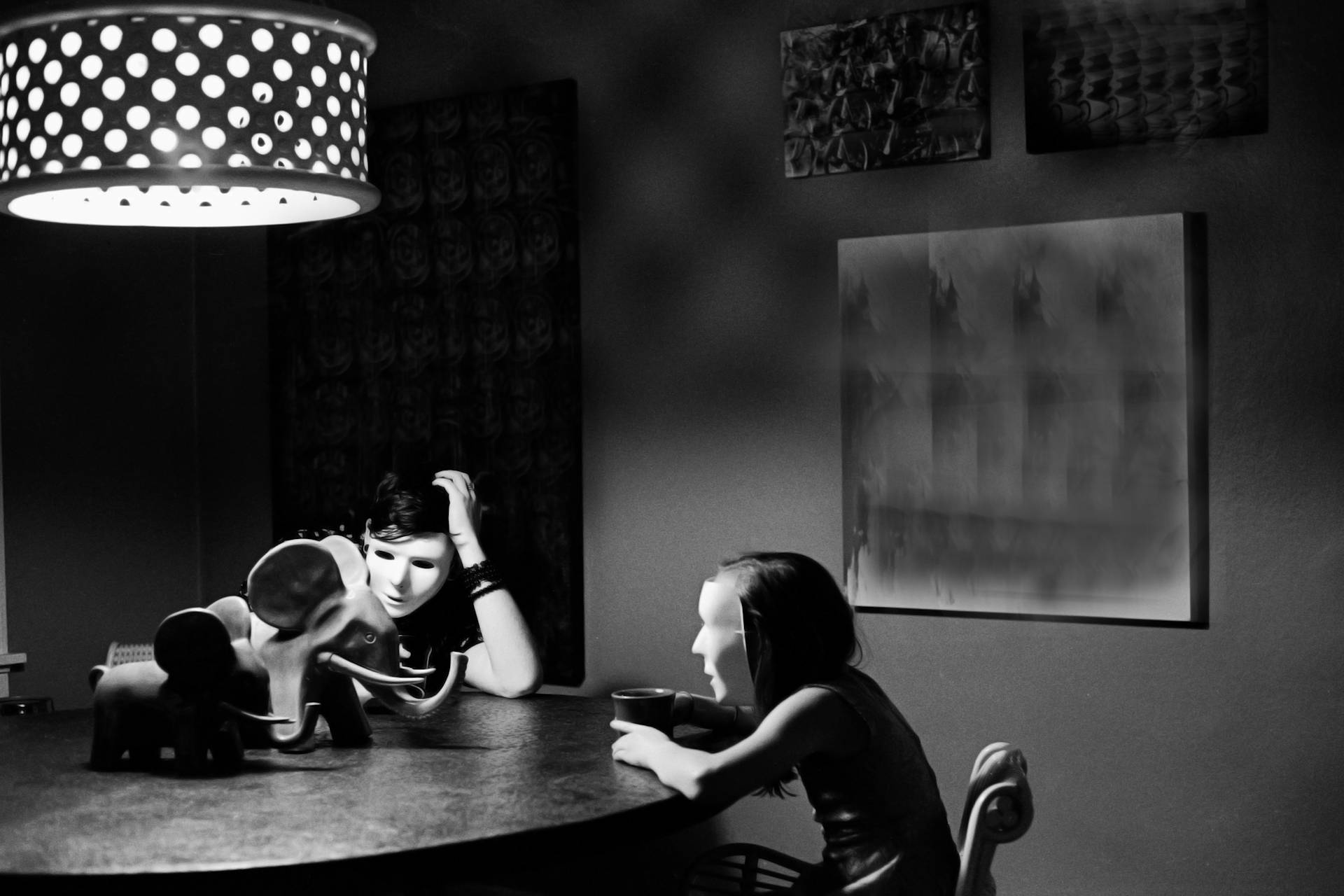 masked children sit slumped at a table looking at a stuffed elephant on the table between them